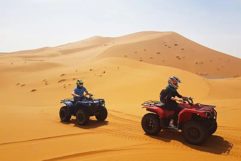 Two riders on quad bikes crossing orange sand dunes in Morocco